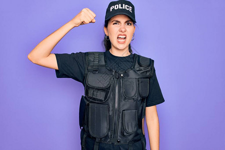 穿着紫色背景的安全防弹背心制服的年轻女警察愤怒地举起拳头图片
