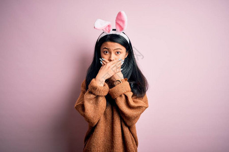 穿着兔子耳朵的年轻女人站在孤立的粉红色背景上用双手遮住嘴顶大惊小怪图片
