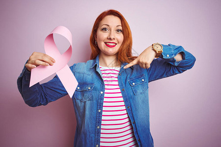 年轻漂亮的红发美女在粉红色孤立的背景上带着癌症丝带并用奇异的图片