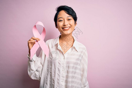 年轻美丽的亚洲女孩在孤立的背景上举着粉色癌症丝带符号图片
