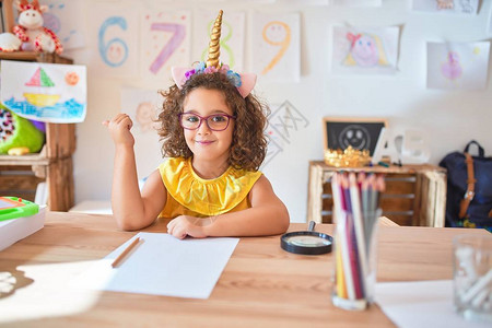 漂亮的蹒跚学步的孩子戴着眼镜和独角兽王冠坐在幼儿园的桌子上图片