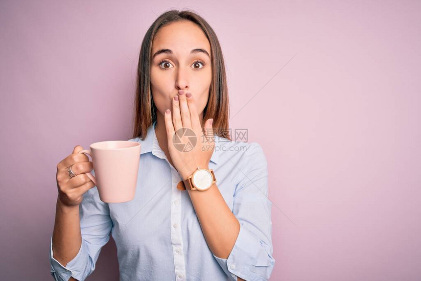 年轻美女喝着咖啡杯站在孤立的粉红背景面罩上手握着满嘴大惊小怪图片