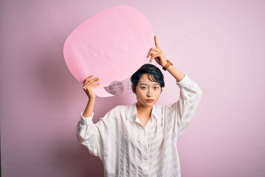 年轻美丽的亚洲女孩在孤立的粉红背景上谈论言语泡沫并且自信地表现在聪明的面孔上图片