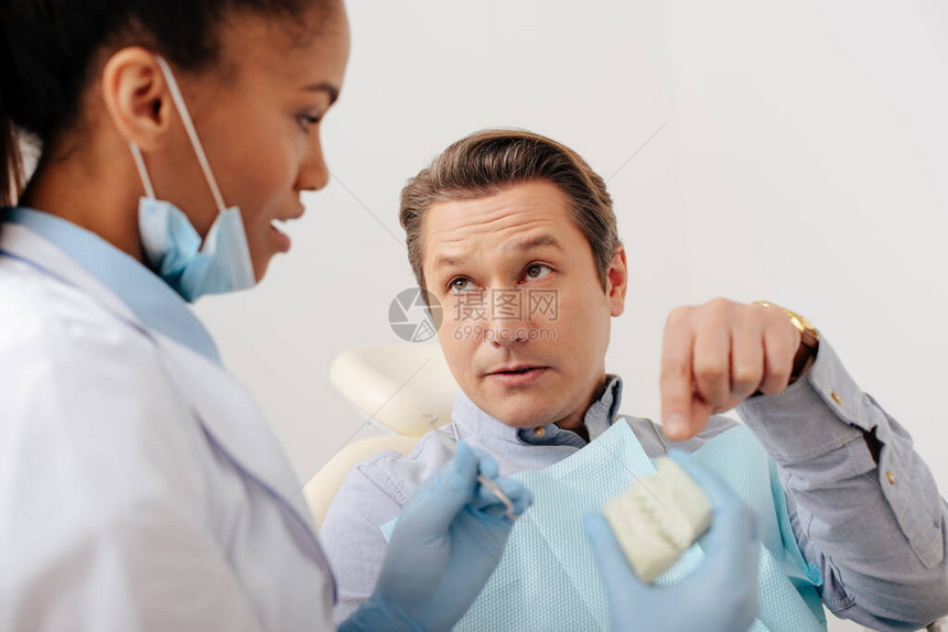 用乳胶手套指着牙型靠近非洲美国牙医的牙模的男人有选图片