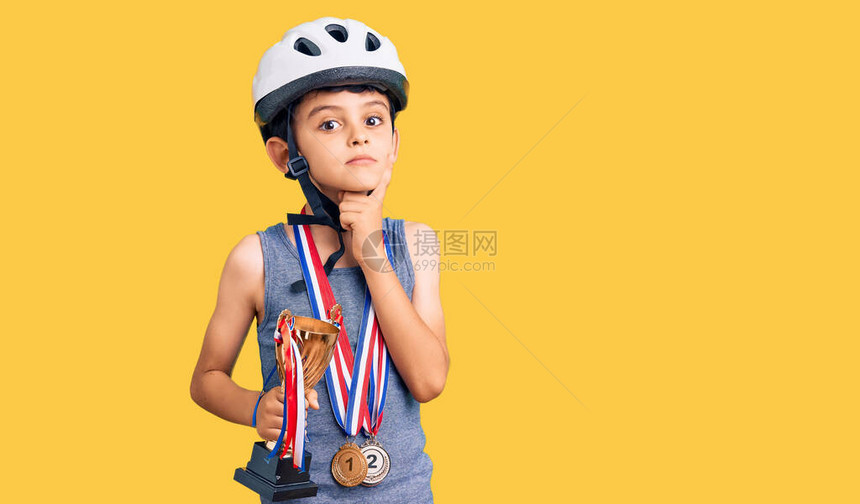 戴着自行车头盔和冠军奖牌的可爱小男孩拿着冠军奖杯严肃的脸用手托着下巴思考问题图片