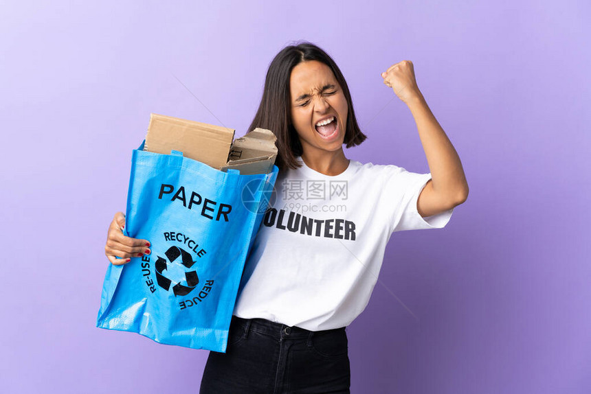 年轻拉廷女人拿着一个回收袋满纸的废纸来回收在紫色上孤立的循图片