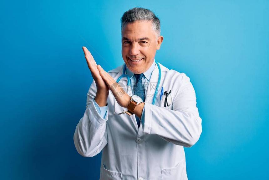 中年英俊的灰发医生穿着外套和蓝色听诊器鼓掌快乐图片