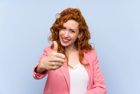 红发女人在孤立的蓝墙上穿着赤发女人用拇指举起手因图片
