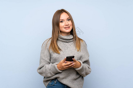 使用手机携带毛衣的青少年女孩在孤立的蓝色背景上图片
