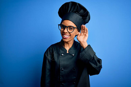 身穿厨具制服戴蓝帽子的年轻非洲女厨师在蓝背景中笑着图片