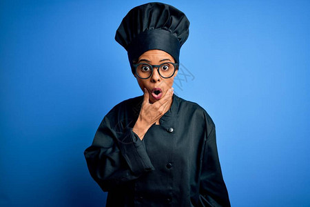 身穿炊具制服和蓝色背景帽子的年轻非洲裔美国厨师女双手托着下巴图片