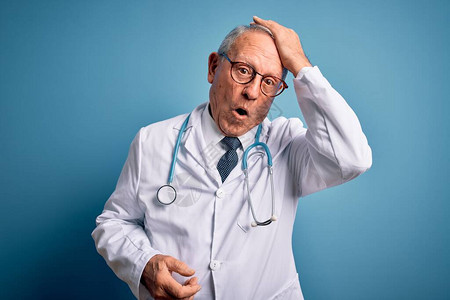 身穿蓝色背景的听诊器和医用外套的白发老医生因错误而惊讶地用手捂着头图片