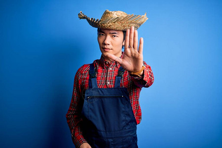 年轻英俊的农民在蓝色背景下穿着围裙和草帽停止用手脸上带着消极和严肃的图片
