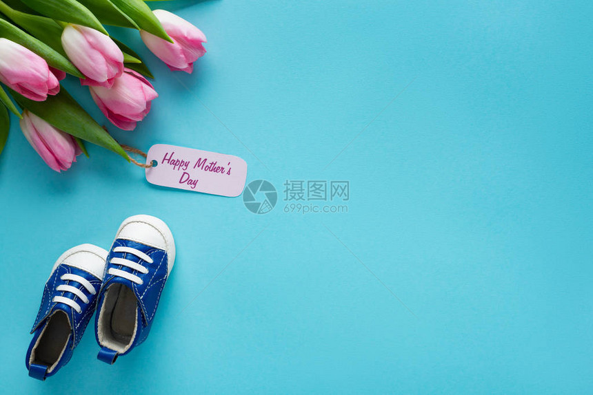 在蓝背景婴儿鞋附近纸贴标签上写字的郁金香和快乐母亲日间喜悦的郁图片