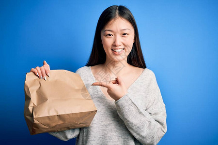 年轻的年青女拿着送纸袋来吃外送食物身穿蓝色孤立背景图片