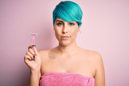 穿着粉色毛巾的蓝发青色时装头发年轻女子拿着美容护理剃刀自信地表现在图片
