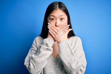 年轻美丽的亚洲女人穿着毛衣站在蓝色的孤立背景上满嘴大惊小怪地用手顶着嘴说错话图片