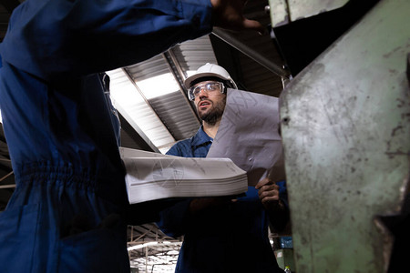 工厂程师阅读机器操作手册并穿着蓝色工作套装和安全帽站立重工业厂概念的技背景图片