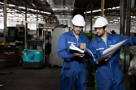 工厂程师阅读机器操作手册并穿着蓝色工作套装和安全帽站立重工业厂概念的技背景图片