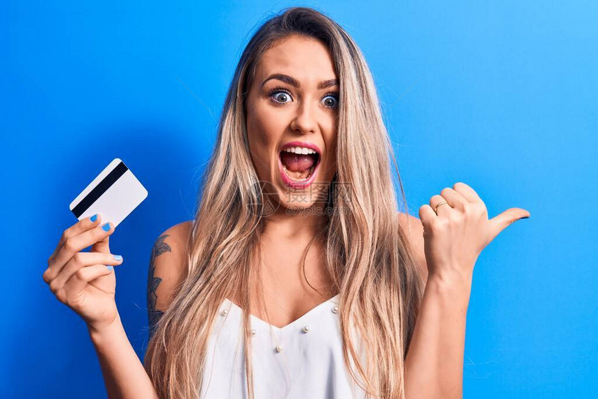 年轻美丽的金发美女拿着信用卡在孤立的蓝背景下举起拇指向侧面用张嘴图片
