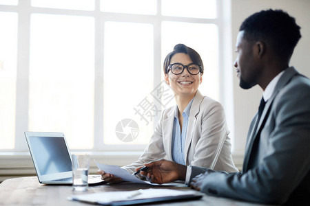 与同事或合作伙伴坐在会议桌旁处理文件时微笑的年轻女商人的肖图片