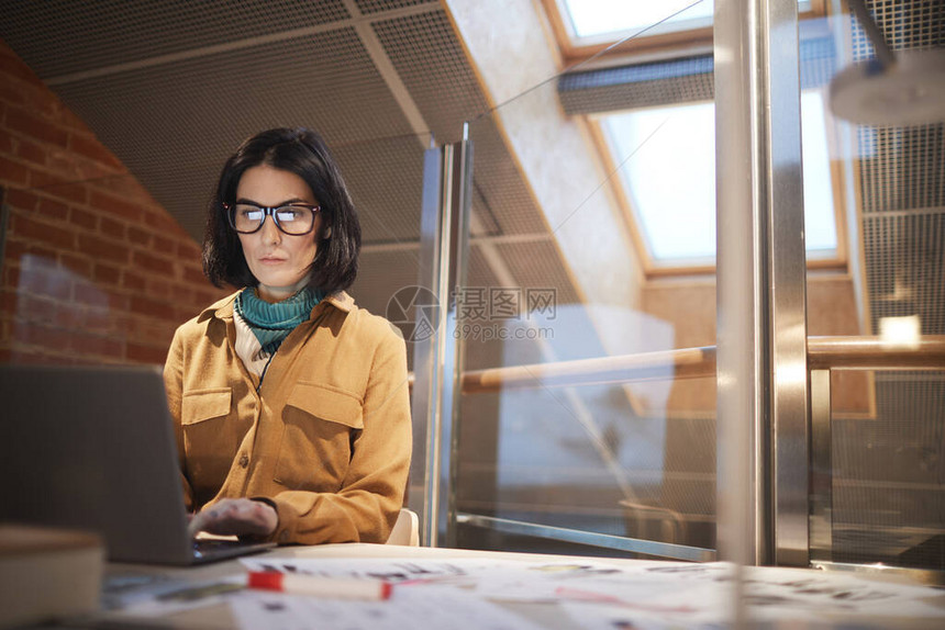 身戴眼镜坐在桌子上并在工作中使用笔记本电脑的图片