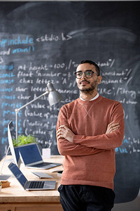 身穿休闲服和眼镜的年轻严肃的交叉武装IT经理站在黑板背景的办公桌旁图片