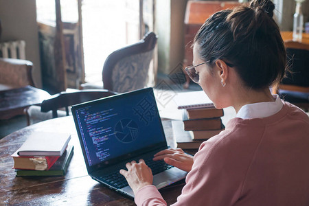 一个戴着黑头发穿粉红色毛衣和眼镜的兴奋女孩坐在图书馆时在笔记本电脑上写代码背景图片