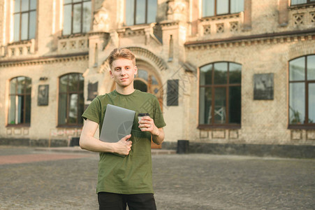 年轻的白人男学生在大学校园前拿着笔记本电脑拿走咖啡图片