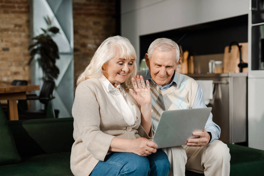 幸福的老年夫妇在自我孤立期间在家里用笔记本电脑挥手和图片