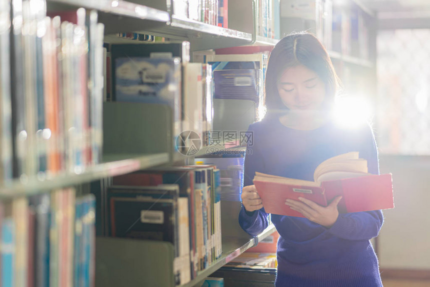 身着休闲西装的亚洲年轻学生站在大学图书馆的书架上阅读书籍或具有各种书籍背景的学院图片