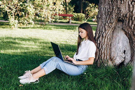 拥有笔记本电脑户外的女学生坐在树旁公园的草地上图片