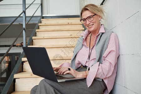 在编制销售报告时坐在楼梯上并使用笔记本电脑的快乐成熟商业图片