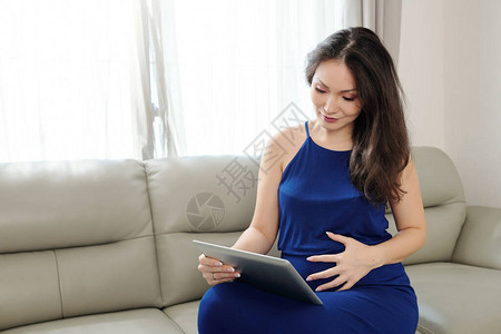 美丽的怀孕年轻女子在平板电脑上观看有关怀孕和分娩的教育视频图片