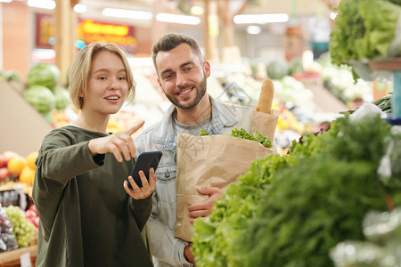 积极的年轻女在市场上购买产品时使用智能手机应用程序购买蔬菜并将图片