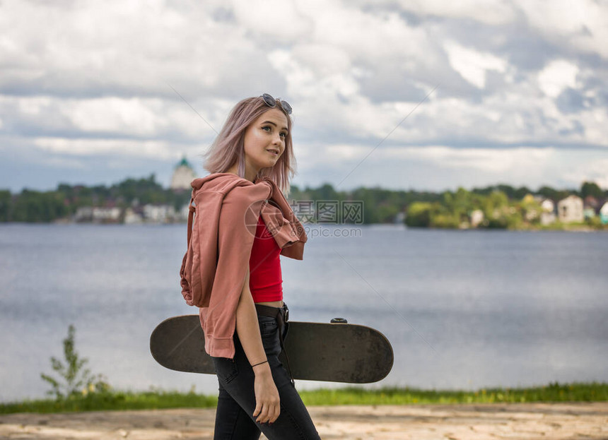 一名带滑板的年轻女子沿着城市池塘的海岸行走图片