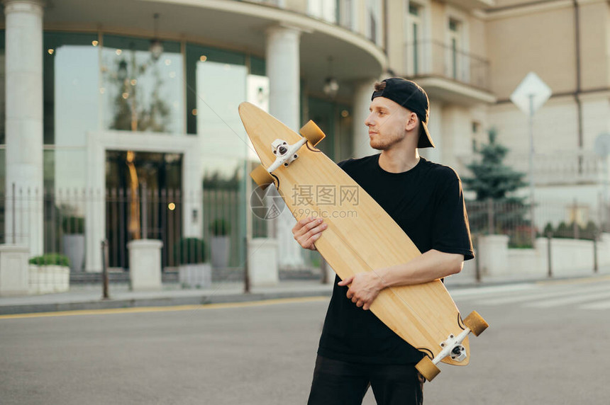 英俊的年轻溜冰者手里拿着一块长板在镇上的街道上摆姿势图片