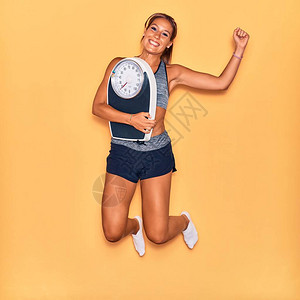 年轻漂亮的西班牙裔女运动员穿着运动服微笑快乐在孤立的黄色背景上拿着称重机的脸上背景图片