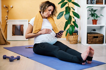 坐在瑜伽垫子上在科罗纳爆发时使用智能手机的中年孕图片