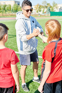 英俊年轻教练与初级足球队在阳光下握手的全长肖像图片