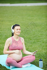 戴着耳机的年轻孕妇坐在运动垫上图片