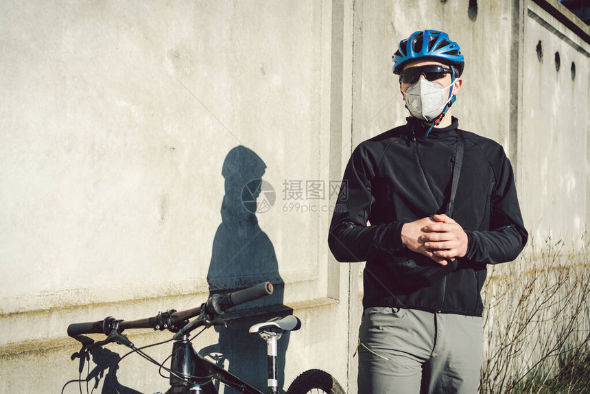 一名戴着防护面罩的男子骑自行车者站在灰色混凝土墙背景的自行车旁边无法辨认的信使在呼吸器中大流行期图片