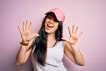 身着随意运动帽的年轻黑发女人在粉红色背景展出和指着十号手指时微笑图片