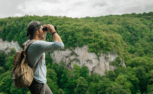 一名男子背着包站在高处使用双筒望远镜检查森林环境研背景图片