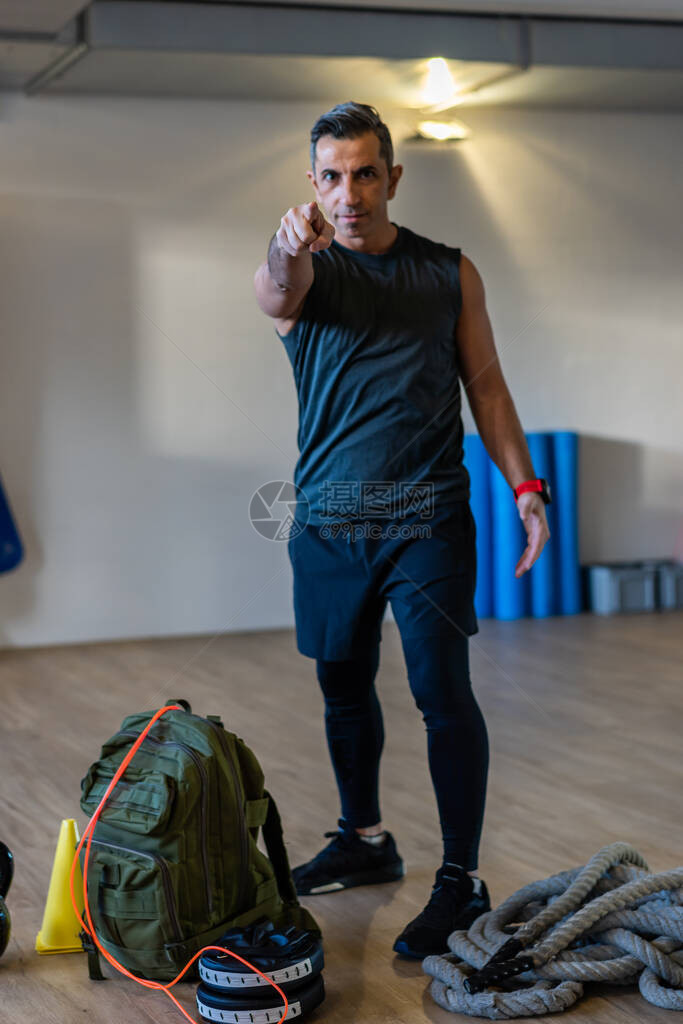 指向照相机的健身教练有动力的新兵训练营教练站在健身房的健身器材上木地板上的哑铃绳索沙袋健身图片