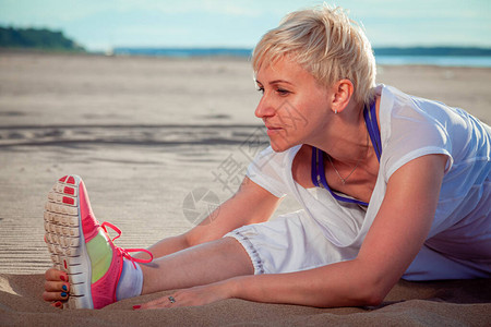 穿着白色运动服的美皮肤金发美女坐在沙滩上做伸展锻炼将她图片