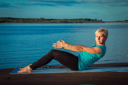 日落时河边沙滩上的蓝地毯上穿着脚踏板的金发女人锻炼身体图片