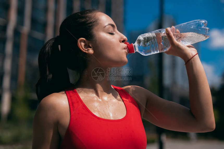 穿着红色胸罩和从塑料瓶里喝水的棕色波浪马尾辫出汗的年轻女子的侧面图背图片