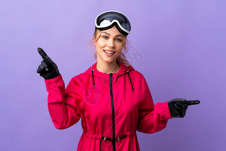 穿滑雪眼镜的滑雪少女在孤立的紫色背景上指着图片