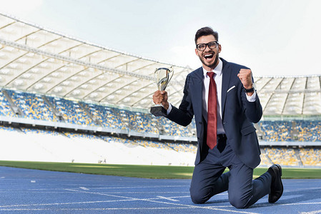 快乐的年轻商人穿着身跪在体育场与奖杯图片